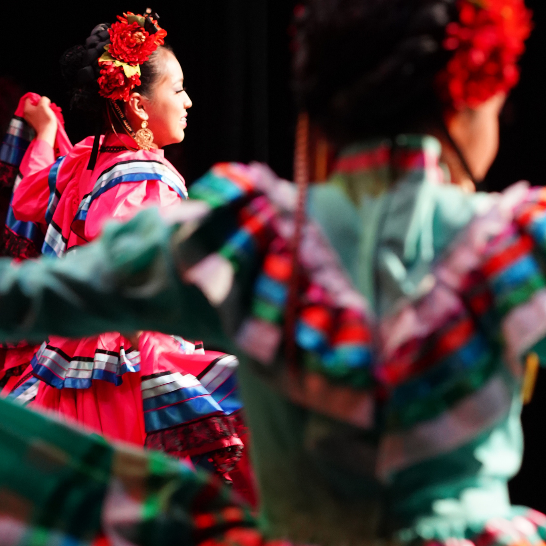 Dancers wearing colorful, layered dresses and floral headpieces perform traditional a Mexican Dance at the Newhall theatre